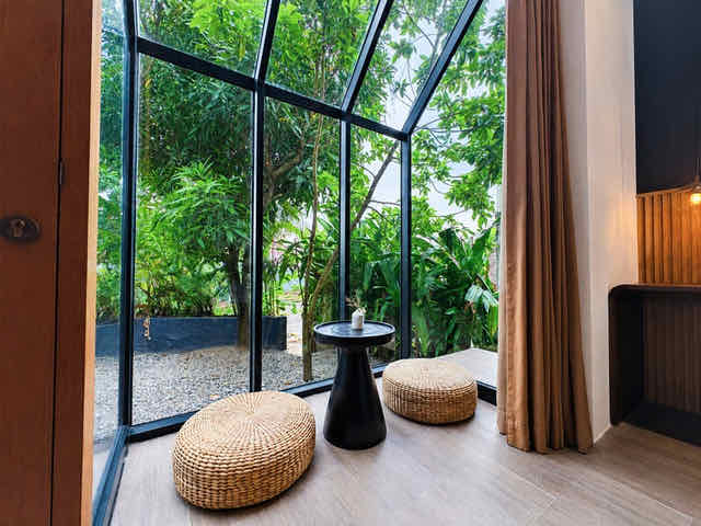 A glass corner of a villa with rattan floor cushions facing the trees